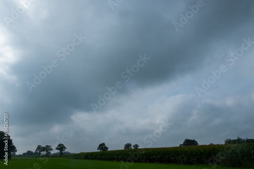 This image depicts a serene countryside landscape with an expanse of green fields under a vast, brooding sky. The ominous clouds gather with a gradient from a lighter grey to a deep, almost black hue