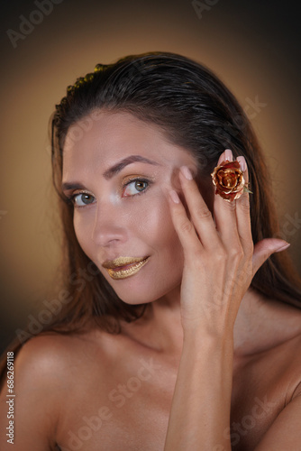 Portrait of brunette female model in studio. Attractive woman with gold lipstick and eyeshadows holding golden rose flower looking at the camera.