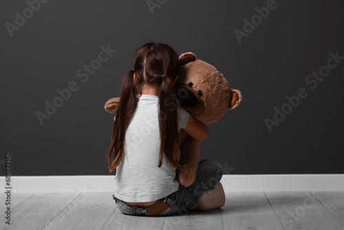 Child abuse. Upset little girl with teddy bear sitting on floor near gray wall indoors, back view © New Africa