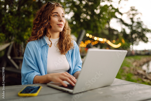 Portrait of a young woman in a blue shirt with a laptop at a table in an outdoor cafe near a lake. Curly woman freelancer works on a laptop outdoors. Freelance concept, nature.