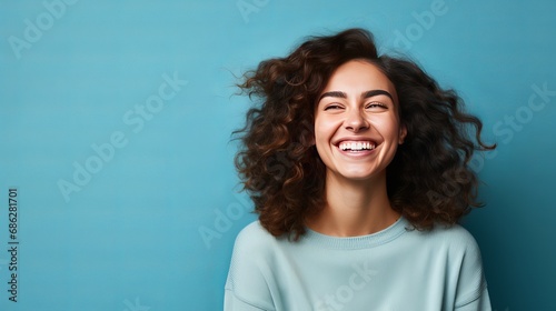 The young woman is laughing joyfully with her arms crossed in a relaxed, positive, and satisfied pose in front of a blue wall.