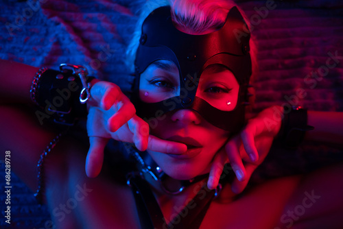 portrait of a sexy girl in a leather mask and bdsm accessories on a bed enjoying sex in orgasm with emotions on her face in neon light photo