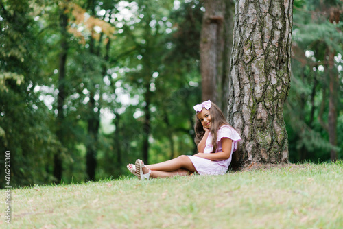 A child girl leans her back against a tree and sits on the grass in summer in an urban landscape park
