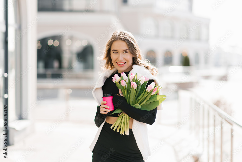 Fototapeta premium Stylish young woman with a bouquet of tulips outdoors and holding a paper cup with coffee in her hands. Portrait of a spring street lifestyle