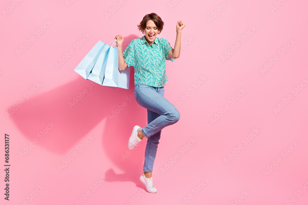 Full body photo of delighted pretty lady hold store bags raise fist empty space isolated on pink color background Full body photo of delighted pretty lady hold store bags raise fist empty space isolated on pink color background