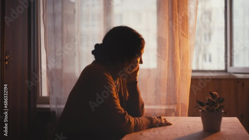 Sad grandmother sitting at table and looking out the window at sunset, nostalgia photo