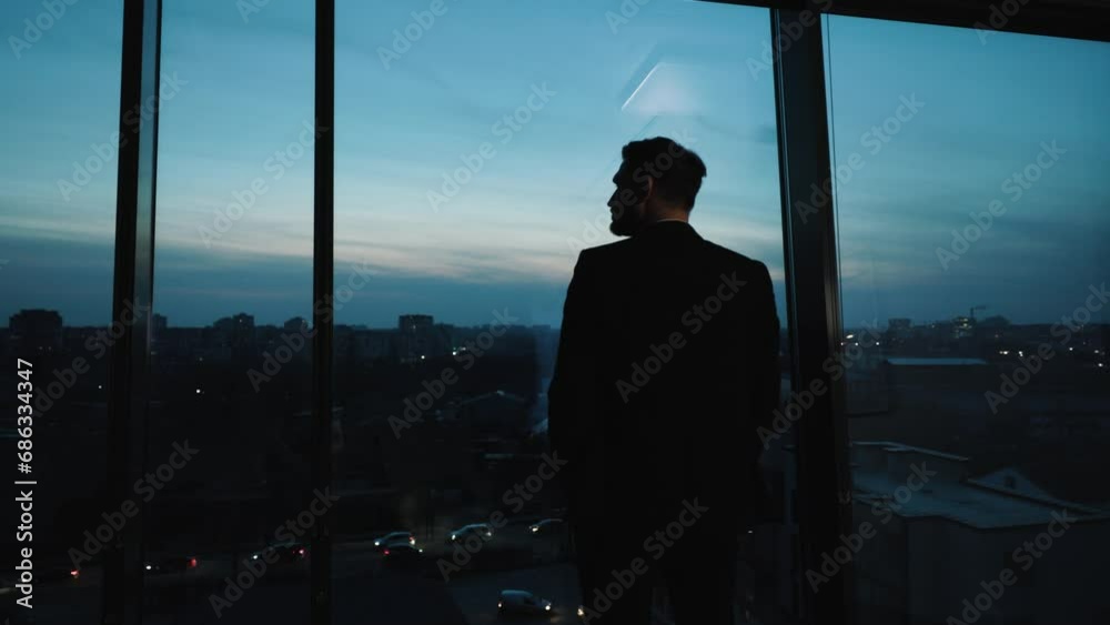 View from behind man in suit standing by panoramic windows in skyscraper, gazing at evening city. Businessman contemplates future while standing in office, observing sunset after completing his work.