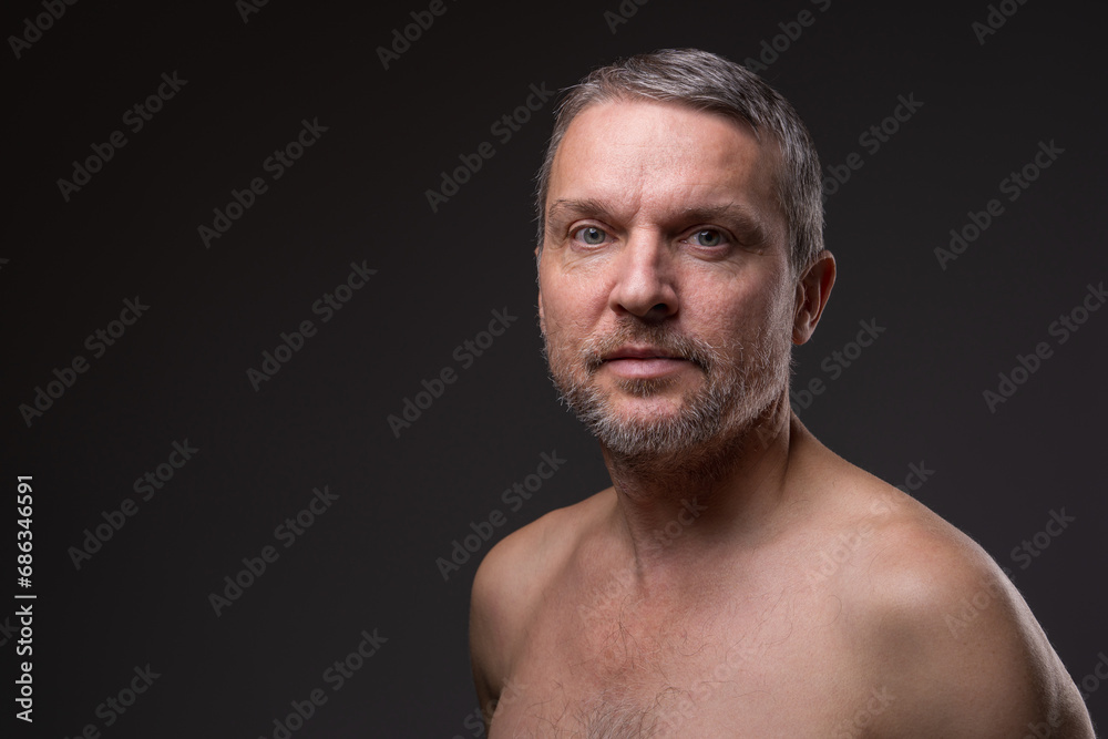 .Slightly smiling face of a middle-aged man with a beard looking at the camera. Close-up portrait of a fifty-year-old man with a naked torso against a dark background. .Slightly smiling face of a middle-aged man with a beard looking at the camera. Close-up portrait of a fifty-year-old man with a naked torso against a dark background.