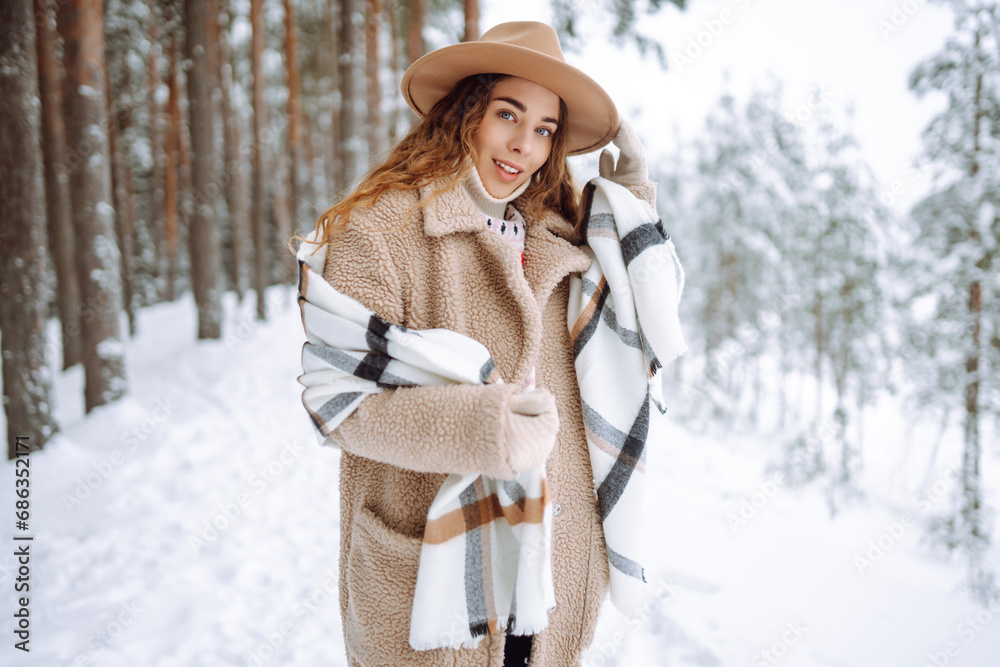 Fototapeta premium Winter walk. A young woman in winter clothes is having fun in a snowy forest. A beautiful female tourist in a hat enjoys a frosty day in the forest.