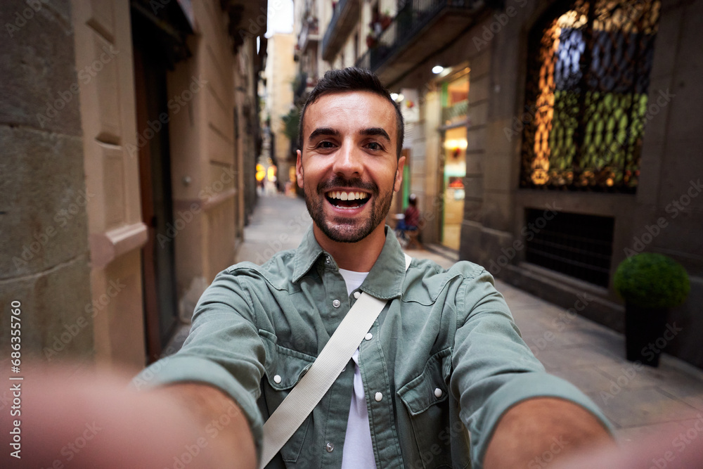 Happy selfie of a young caucasian man in an old town of Barcelona. Male tourist taking a self portrait using smartphone to post it on social media, - obrazy, fototapety, plakaty 