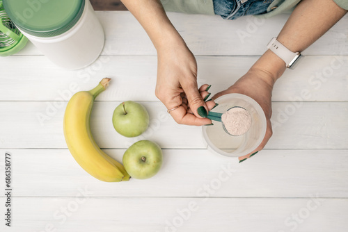 Young woman with measuring spoon in her hand puts portion of whey protein powder into shaker on white wooden table with bananas and apples. Process of making protein drink, top view