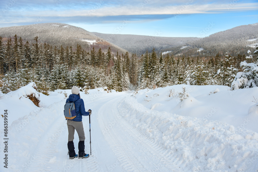 A young woman is standing on a snow-covered hiking trail and admiring the panorama of the Sudetes mountains.