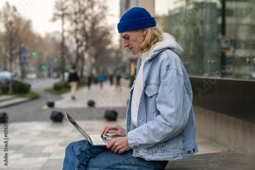 Focused man with laptop sits near building works as programmer in IT company outdoors. Carried away blond guy works as freelancer in big tech industry for corporations or startups. Distance work