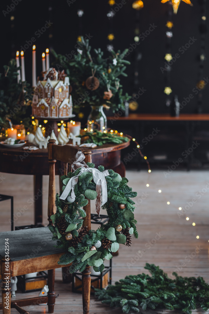Naklejka premium Christmas elegant composition with handmade wreath on wooden chain and beautiful homemade gingerbread house, candles on the table. Family party festive decoration, cozy atmosphere, aesthetics