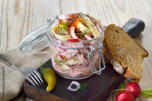 Deftiger Schweizer Wurstsalat mit Käsestreifen und Essiggurken im Glas, dazu rustikales Bauernbrot – Hearty sausage salad with cheese strips and pickles in a glass jar, served with  farmhouse bread photo