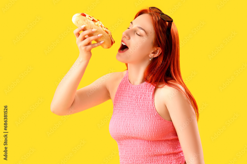 Beautiful young woman eating tasty hot dog on yellow background Beautiful young woman eating tasty hot dog on yellow background