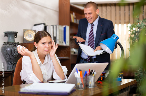 Young businessman who is in the office scolds his subordinate girl sitting at workdesk, pointing out her mistakes