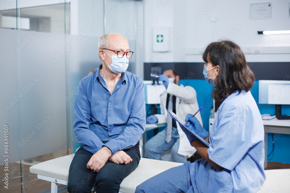 Nurse practitioner is taking notes during a session with senior man. During covid 19 epidemic, an elderly male patient attending a healthcare assessment with a medical assistant. Nurse practitioner is taking notes during a session with senior man. During covid 19 epidemic, an elderly male patient attending a healthcare assessment with a medical assistant.