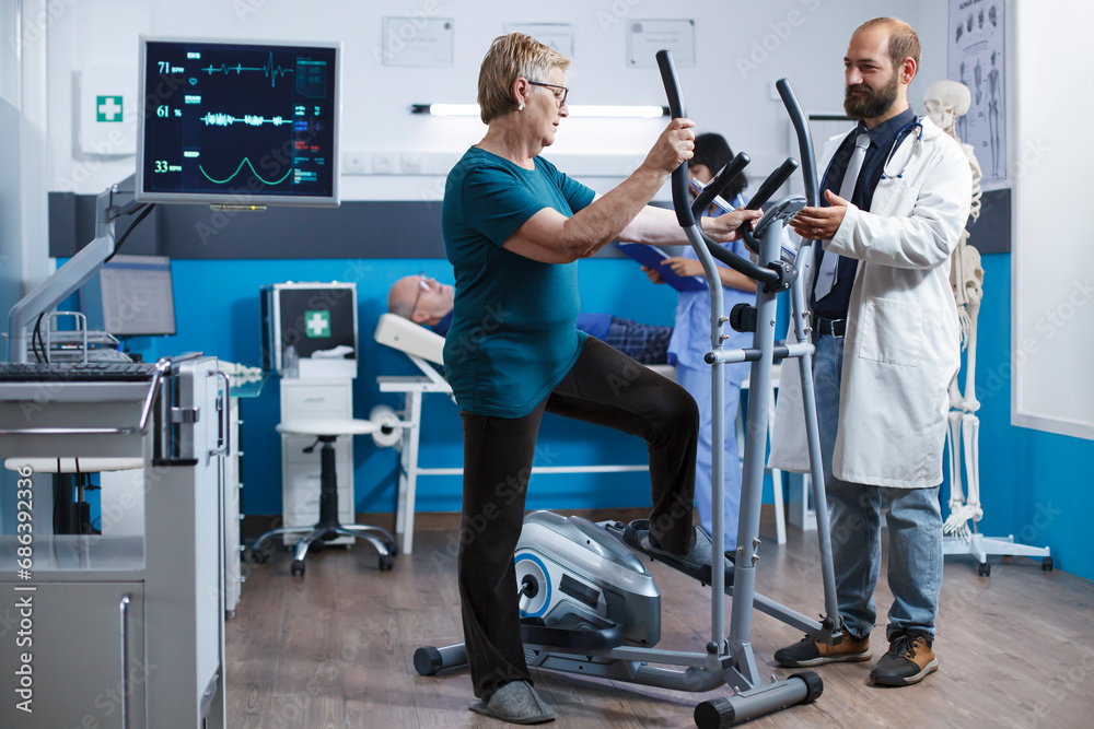 Elderly woman uses electric bicycle for physical rehabilitation after being hurt. Senior female patient using a stationary bike for gymnastics while the doctor assists with physiotherapy. Elderly woman uses electric bicycle for physical rehabilitation after being hurt. Senior female patient using a stationary bike for gymnastics while the doctor assists with physiotherapy.