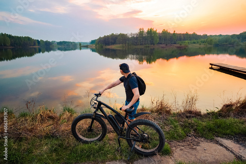 Man with an electric fatbike on the background of a lake and sunset. Picturesque place in the village. Concept of a healthy lifestyle.