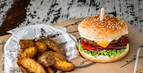 Beef burger with lettuce, tomatoes, slice of cheese, pickles, sauce, onions rings, french fries on vintage newspapper over wooden background, close up photo