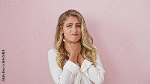 Young blonde lady in pain, clutching her sore throat as flu and infection symptoms show, standing against an isolated pink background, with a heartbroken expression photo
