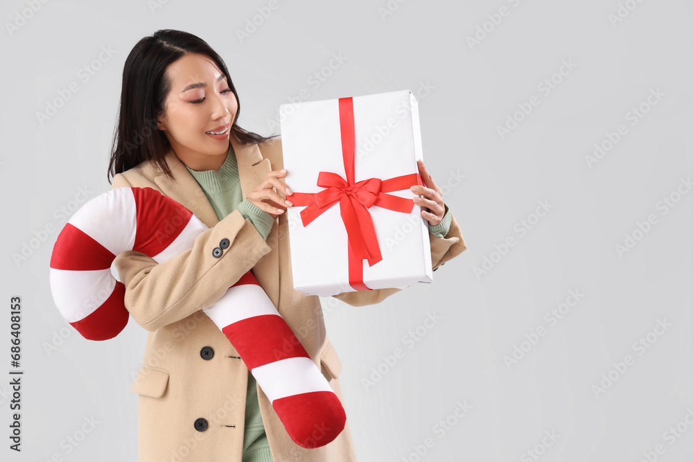 Young Asian woman with Christmas gift and candy cane pillow on light background Young Asian woman with Christmas gift and candy cane pillow on light background