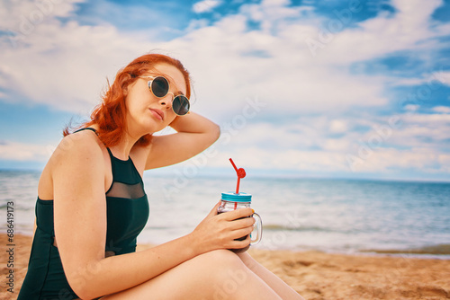 Beautiful woman in swimsuit is sitting on beach with cocktail and look away. Tropical alcohol beverage. Red-haired lady with sunglasses on vacation. Summer day with cloudy sky. Holidays at sea.