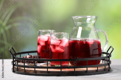 Refreshing hibiscus tea with ice cubes and dry roselle flowers on white wooden table against blurred green background