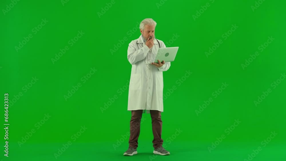 Portrait of aged man medic on chroma key green screen. Senior doctor in white coat standing typing on laptop, reading information online. Portrait of aged man medic on chroma key green screen. Senior doctor in white coat standing typing on laptop, reading information online.