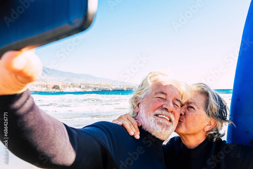 beautiful couple of two seniors at the beach with wetsuits and surfboard taking a selfie before go surf together - active mature people photo