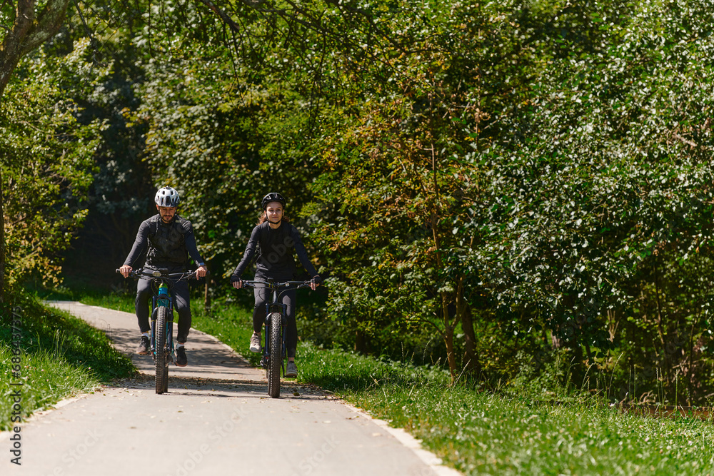Obraz premium A blissful couple, adorned in professional cycling gear, enjoys a romantic bicycle ride through a park, surrounded by modern natural attractions, radiating love and happiness