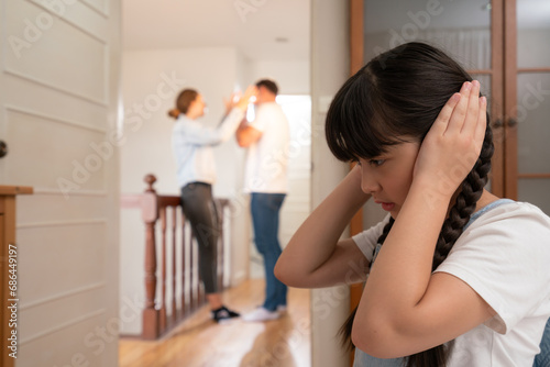 Stressed and unhappy young girl huddle in corner, cover her ears blocking sound of her parent arguing in background. Domestic violence at home and traumatic childhood develop to depression. Synchronos