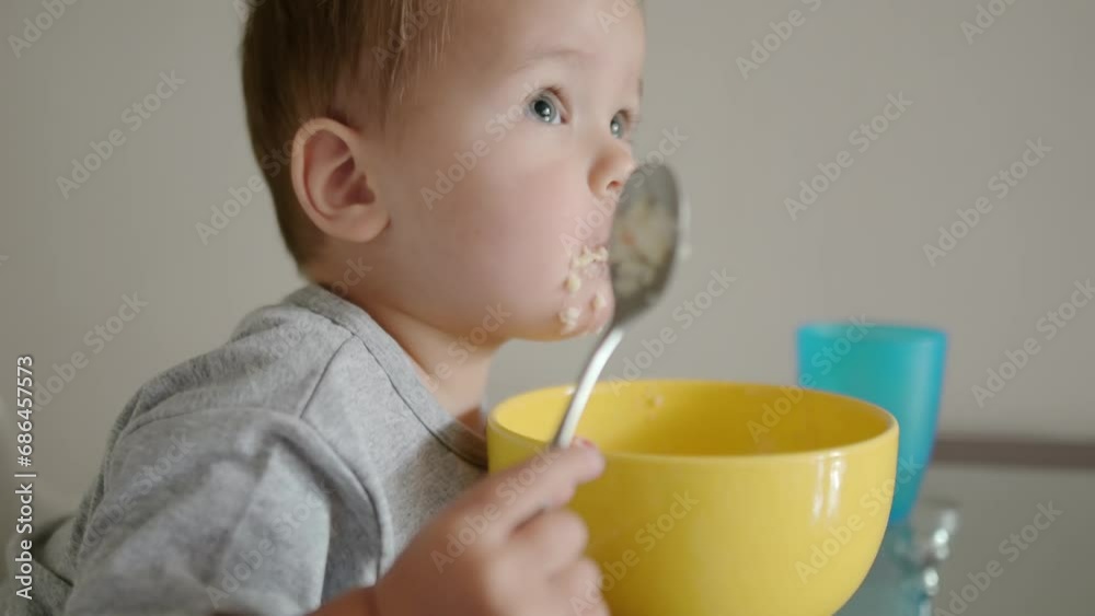 A cute toddler boy eating a healthy breakfast of cereal in the kitchen in the morning. A baby learning to eat with a spoon. Complementary feeding are new stage of feeding of child. A cute toddler boy eating a healthy breakfast of cereal in the kitchen in the morning. A baby learning to eat with a spoon. Complementary feeding are new stage of feeding of child.