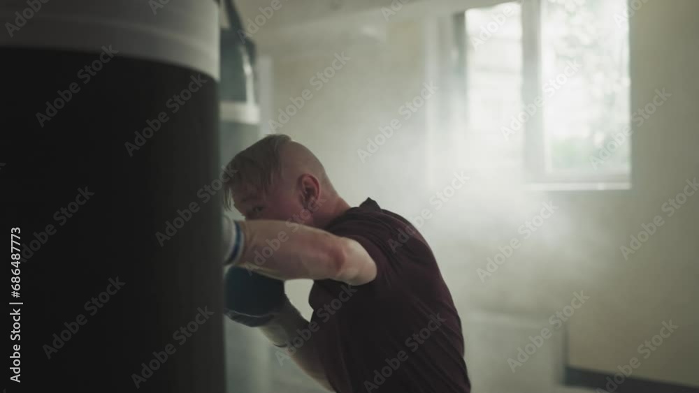 Sportsman punches boxing bag in sports center. Joyful athlete in gloves beats hanging equipment by arms in smoky gym slow motion. Combat attack style Sportsman punches boxing bag in sports center. Joyful athlete in gloves beats hanging equipment by arms in smoky gym slow motion. Combat attack style