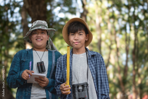 Young asian schoolboy is standing in school botanical garden and measuring a size of tree trunk with a measuring tape and recording information of trees for school botanical garden library.