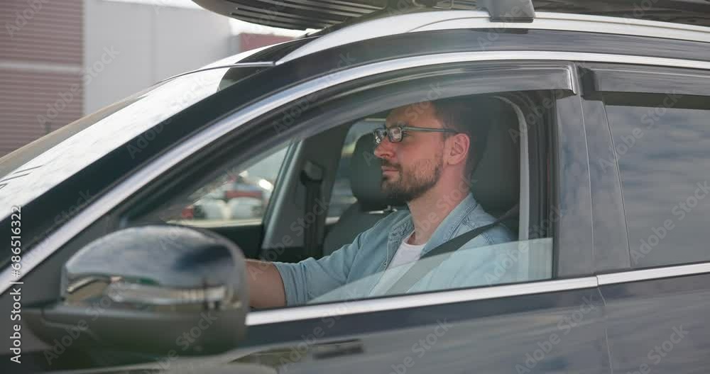 Young Caucasian man sits in a car on a parking lot and looking at camera through an open window Young Caucasian man sits in a car on a parking lot and looking at camera through an open window