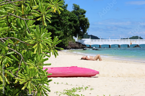 View to tropical sandy beach through leaves of Scaevola taccada. Woman tanning on sea coast, vacation and travel