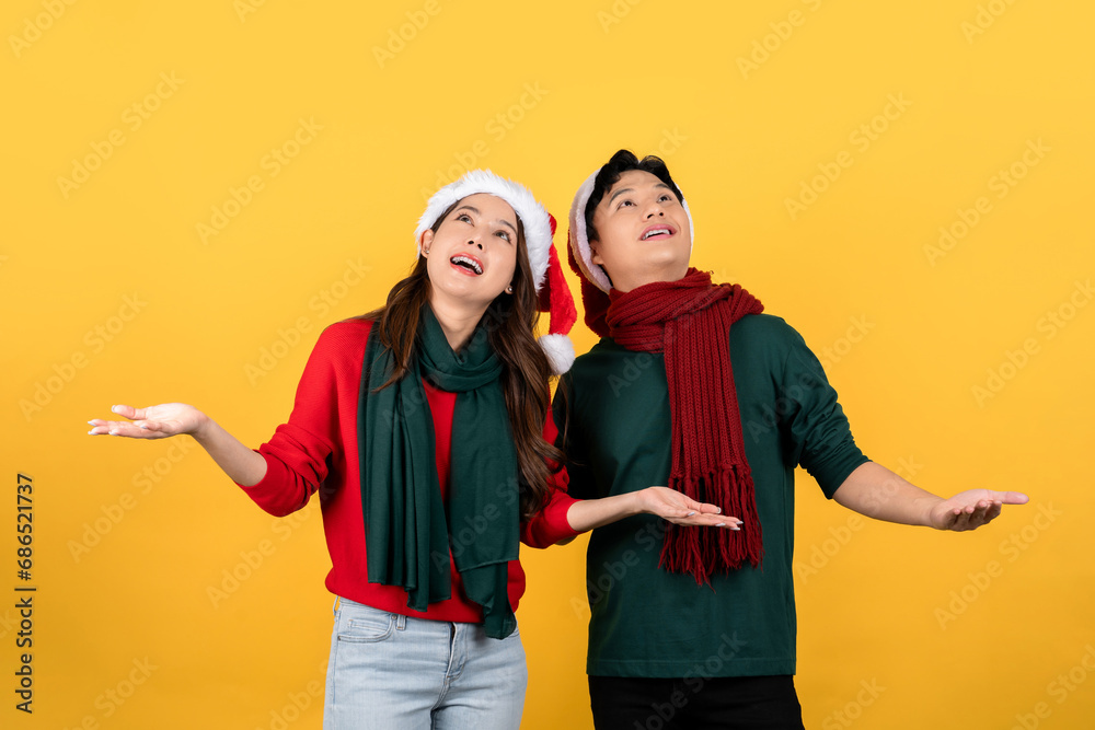 Obraz premium Photo of an excited Asian couple in Christmas clothes looking up with open hands on a yellow studio background.