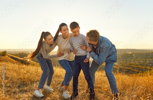 Happy smiling family of four with two kids boy and girl having fun and walking outdoors enjoying beautiful nature. Young joyful parents with children hugging spending time together at sunset.