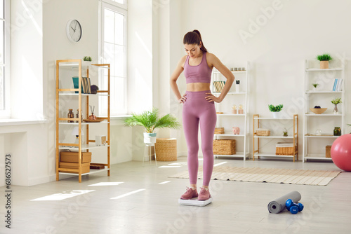 Beautiful fit slim young woman in sportswear standing with her hands on hips on scales in living room and looking down at numbers. Concept of fitness, weight control, weight loss, and sports workouts