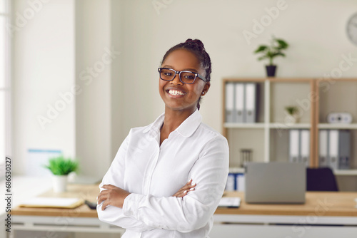 Portrait of successful, proud and positive African American business woman posing in office. Happy dark-skinned woman in white shirt and glasses stands with her arms crossed and smiles at camera.