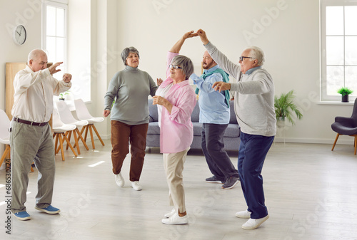Retirement community. Elderly men and women actively spend time having fun and dancing at party in nursing home. Group of senior Caucasian people laughing and dancing together in living room.