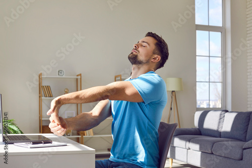 Working man takes a break for stretching exercise. Man in blue T shirt sitting at desk with laptop computer at home and doing relaxing moves to prevent muscle pain and discomfort in arms and shoulders photo