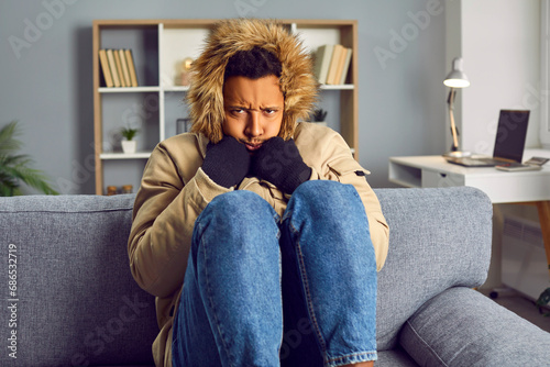 Portrait of a young angry african american frozen man sitting in a warm winter coat with a hood and black gloves on the grey sofa at home and trying to keep warm himself. Heating problems concept.