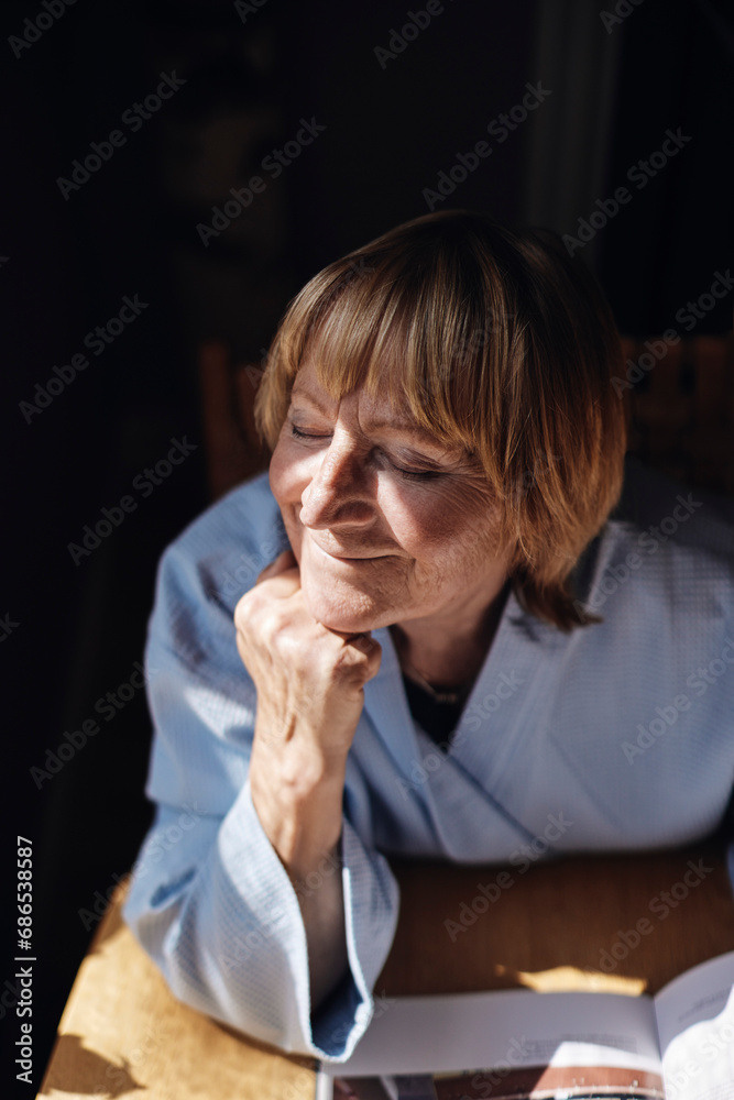 Blond senior woman with hand on chin relaxing in sunlight at home Blond senior woman with hand on chin relaxing in sunlight at home