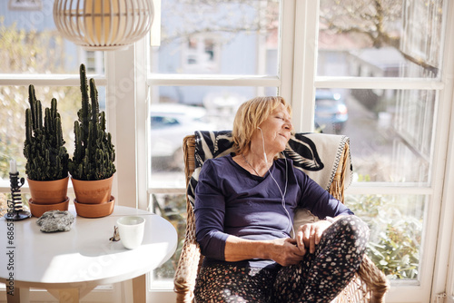 Senior woman with eyes closed listening music through in-ear headphones while relaxing on chair at home