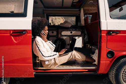 Side view of young woman talking on smart phone while using laptop sitting in van during vacation photo