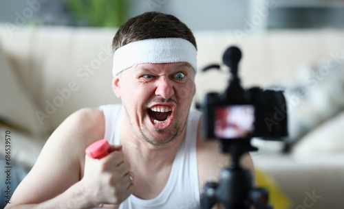 Portrait of an emotionally male blogger with grimace on face and holding dumbbells in front of camera. Sloth fatigue and daily sports training photo