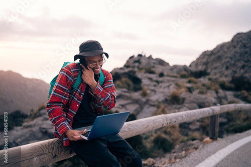 Smiling male explorer talking on smart phone while sitting with laptop on railing photo